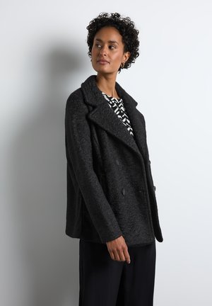 Black double-breasted coat with a textured finish, wide lapels, and large buttons. Worn over a black-and-white patterned shirt.