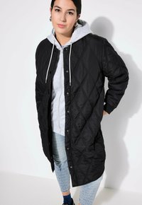 Cappotto lungo imbottito nero con motivo a diamante, dotato di cappuccio, bottoni a scatto sul davanti e polsini a coste. Indossato sopra un hoodie grigio e jeans.