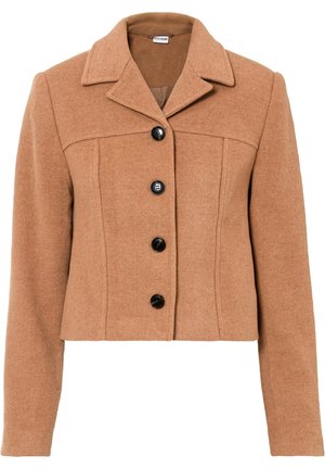 Donna che cammina indossando una giacca corta color cammello, pantaloni neri e stivaletti neri con tacco su sfondo bianco.