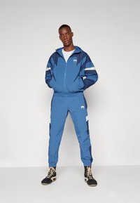 Blauwe trainingsoutfit gemaakt van lichtgewicht stof, met een zip-up jack met panelen, elastische boorden en bijpassende broek met zijaccenten.