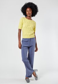 Gelber Strickpullover mit einem strukturierten Wellenmuster, kombiniert mit hellblauen, maßgeschneiderten Hosen und silberakzentuierten Schuhen.