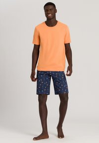 T-shirt arancione a maniche corte abbinato a pantaloni da bagno blu navy con motivi a onde e disegni circolari. Modello a piedi nudi, in posizione eretta.