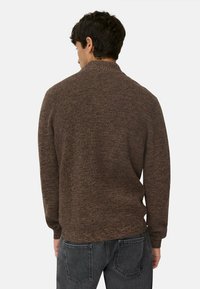 Brauner strukturierter Pullover mit hohem Kragen, langen Ärmeln und taillierter Silhouette, kombiniert mit grauen Jeans.