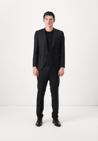Completo nero a righe con giacca aderente e pantaloni abbinati, indossato sopra una maglietta nera, abbinato a scarpe eleganti nere.