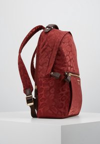 Punainen nailoninen reppu, jossa on s ävy sävyyn leopardikuosi, ruskeat yksityiskohdat ja kultaiset metalliosat. Varustettu pehmustetuilla olkahihnoilla ja vetoketjullisella etutaskulla.