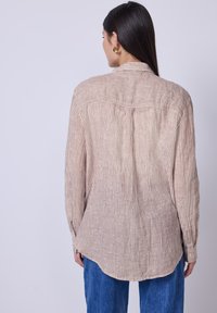 Donna con lunghi capelli scuri che indossa orecchini a cerchio dorati, una maglietta a maniche lunghe a righe beige e jeans blu, vista da dietro su uno sfondo neutro.