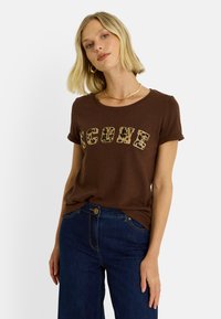 Camiseta corta de manga corta color marrón con cuello redondo, con la palabra "ICON" en letras grandes con estampado de leopardo en color beige. Combinada con jeans de lavado oscuro.