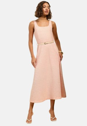 Vrouw met een mouwloze, gestructureerde roze midi-jurk met een dunne beige riem, gouden armband en witte sandalen met bandjes, staand tegen een witte achtergrond.