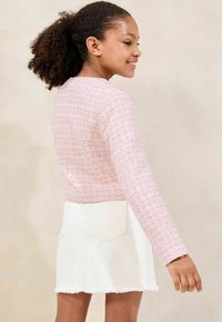 Hellrosa strukturierte Strickjacke mit langen Ärmeln, kombiniert mit einem weißen Denim-Minirock, der über zwei Gesäßtaschen und einen fransigen Saum verfügt.
