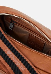 Interno di una borsa in pelle marrone con tasca con zip, fodera testurizzata, tracolla nera con motivo a cuori e un'etichetta in pelle con marchio.