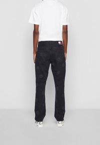 Black denim jeans with smiley face embroidery throughout, featuring a straight-leg cut and a logo patch on the waist. Paired with white sneakers.