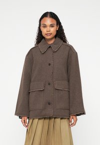 Bruine visgraatjas met een rafelige kraag en zoom, voorzien van twee voorzakken en zwarte knopen, gedragen over een geplooide rok.