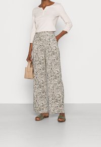 Hellrosa Langarm-Henley-Top, weite Blumenhose in Beige, olivgrüne flache Sandalen und eine kleine gewebte Handtasche in Beige.