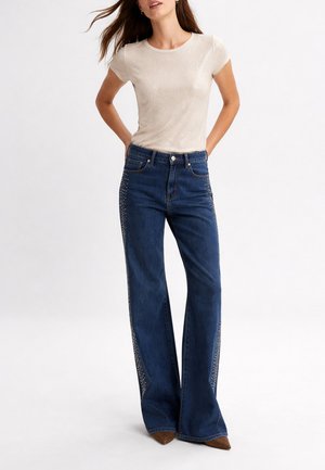 Donna in piedi con le mani dietro la schiena, indossa una maglietta beige a maniche corte e jeans blu scuro a vita alta con dettagli in strass lungo i lati.