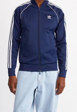 Mężczyzna ubrany w granatową kurtkę dresową Adidas z białymi paskami na rękawach oraz jasnoniebieskie dżinsy, stojący na tle białego tła.