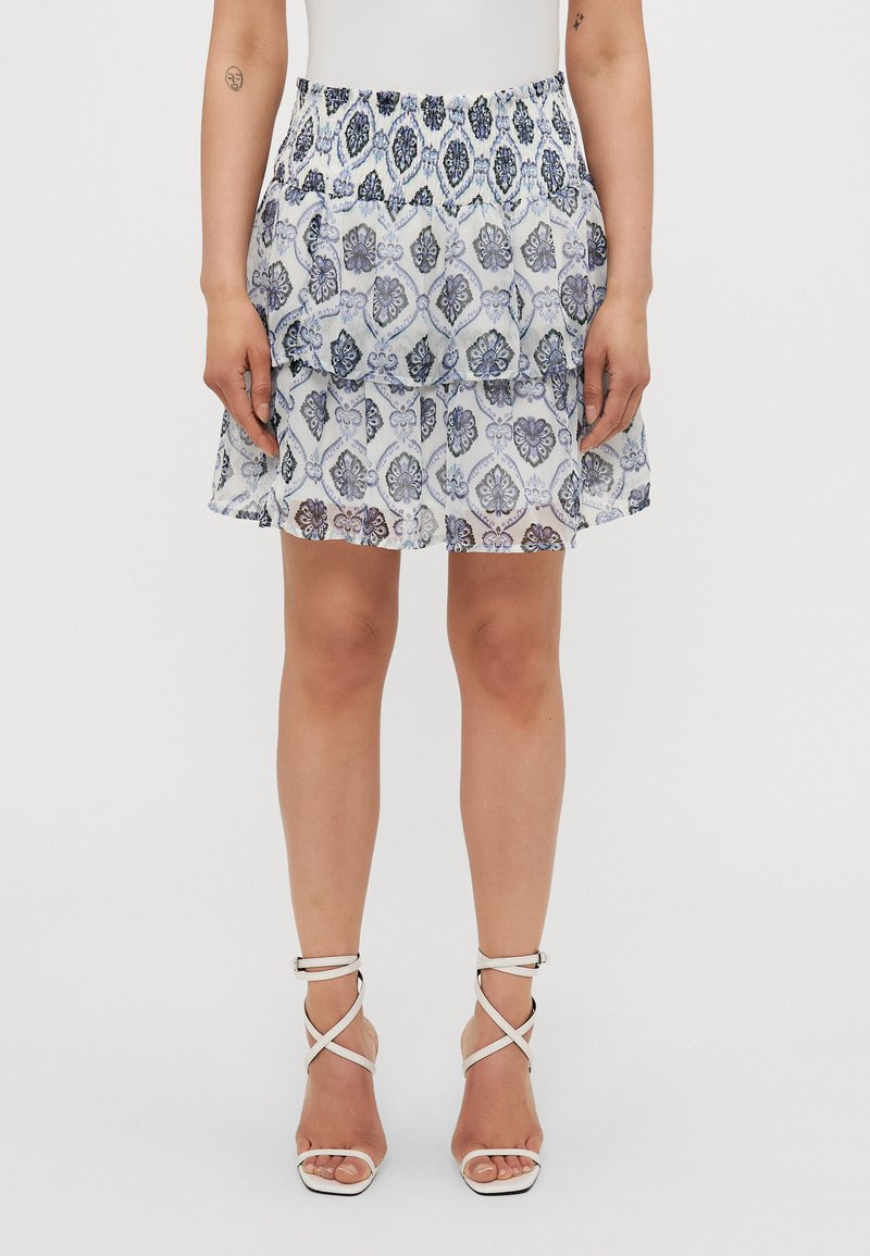 Falda blanca con estampado floral en azul y púrpura, cinturilla fruncida, diseño en capas y detalles con volantes. Usada con tacones blancos de tiras.