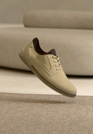 Chaussure beige à lacets avec doublure intérieure marron foncé semblant flotter au-dessus d’un tapis texturé dans une pièce minimaliste, doucement éclairée aux tons neutres.