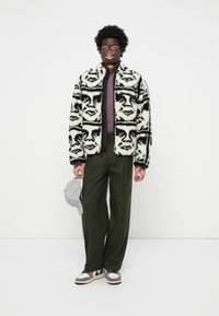 Chaqueta de forro polar con patrones de caras en blanco y negro, con cremallera al frente, combinada con pantalones de color verde oliva y una gorra gris claro. Las zapatillas completan el conjunto.