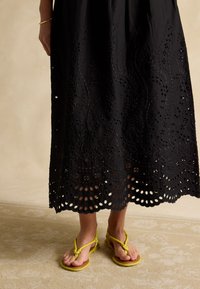 Lower legs in yellow strappy sandals, wearing a black midi skirt with floral embroidery and scalloped eyelet hem.