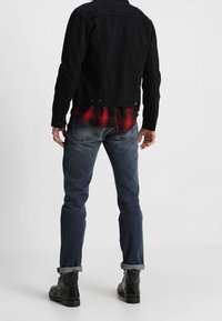 Svart denimjacka över en röd och svart rutiga flanellskjorta, blåjeans och svarta stövlar. Medelblå jeans med uppvikta ärmar och synlig söm.