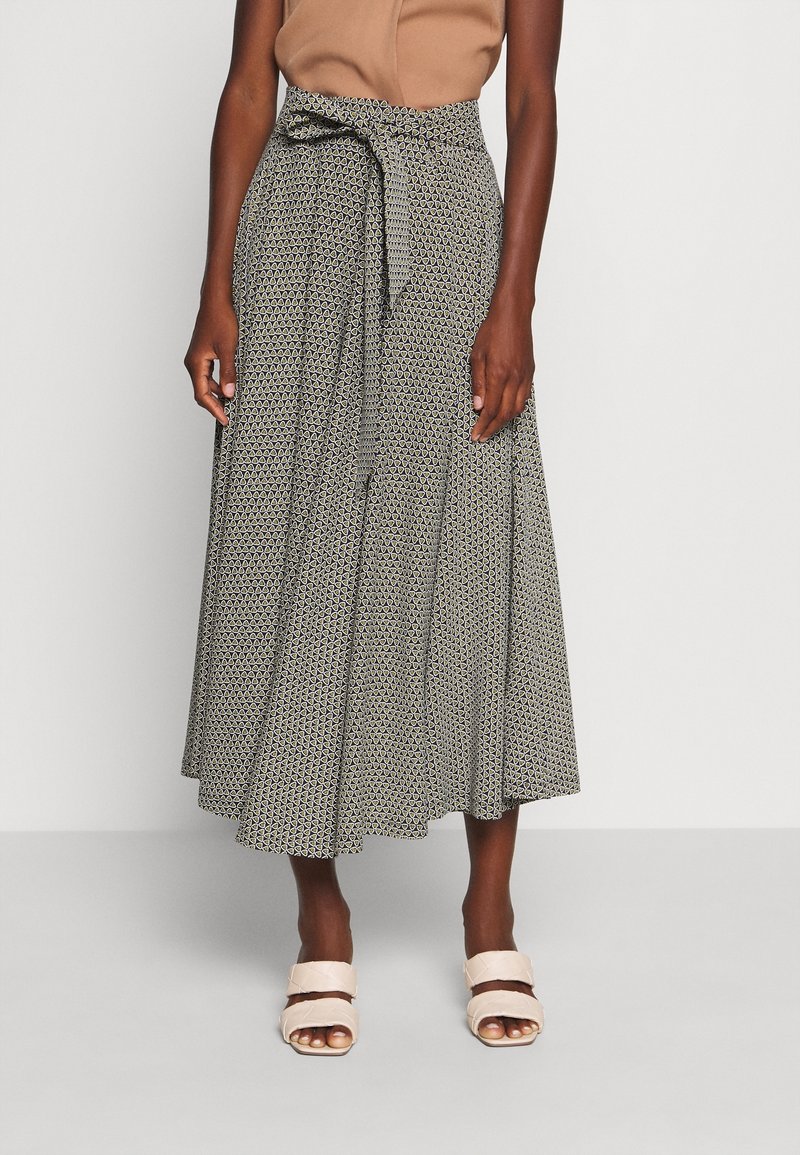 Femme portant une jupe midi à motifs taille haute avec une ceinture nouée, un top sans manches beige et des sandales à talons ouverts beiges.