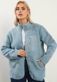 Giacca in pile blu cielo con colletto alto, superficie testurizzata e tasca frontale con zip in un materiale a contrasto. Maniche lunghe e tasche laterali.