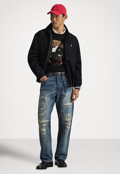 Black zip-up jacket, graphic sweater with teddy bear design, light blue distressed jeans, red cap, and black shoes. Simple accessories visible.