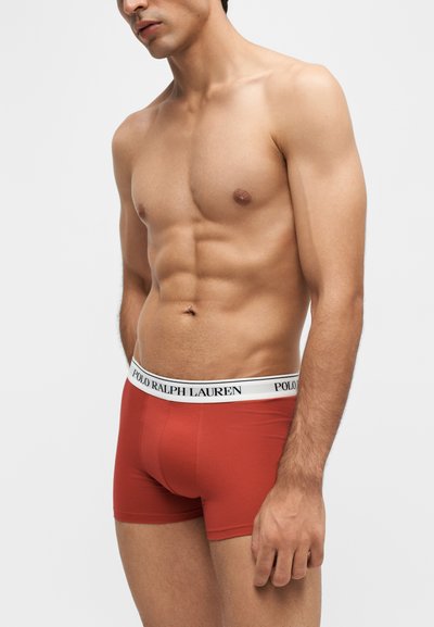 Μυώδης άντρας που φοράει κόκκινα boxer Polo Ralph Lauren με λευκή ζώνη με λογότυπο, στέκεται μπροστά σε ένα απλό φωτεινό φόντο.