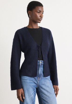 Marineblau, flauschiger Strickcardigan mit V-Ausschnitt und offener Vorderseite. Verfügt über lange Ärmel und eine Bindeschlaufe in der Taille, kombiniert mit hellblauen Jeans.