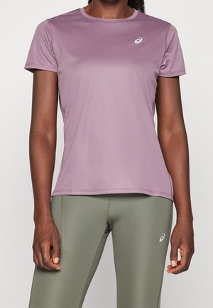 Kvinde iført en kortærmet, lyngfarvet sportstop og olivengrønne leggings med sportsbrandlogoer på brystet og låret.