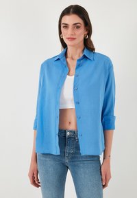 Camicia azzurra chiara in un materiale liscio, con colletto classico, maniche arrotolate e orlo dritto.
