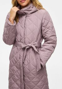 Žena v mauve prešívanom kabáte s opaskom zviazaným do mašle, jedna ruka pri golieri a druhá v vrecku.