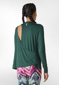 Top verde a maniche lunghe con schiena aperta, caratterizzato da un design drappeggiato e una morbida texture. Abbinato a leggings a zigzag multicolori.