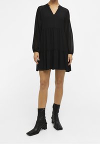 Robe noire à volants avec un décolleté en V, des manches longues en tissu transparent, et une silhouette fluide. Associée à des bottines en cuir noires à bout carré.