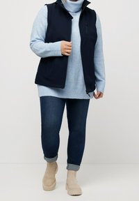 Marineblauer ärmelloser Pullover über einem hellblauen Rollkragen, kombiniert mit einer dunklen Jeans und beigen Stiefeletten. Mit gerippten Akzenten und Reißverschlüssen.