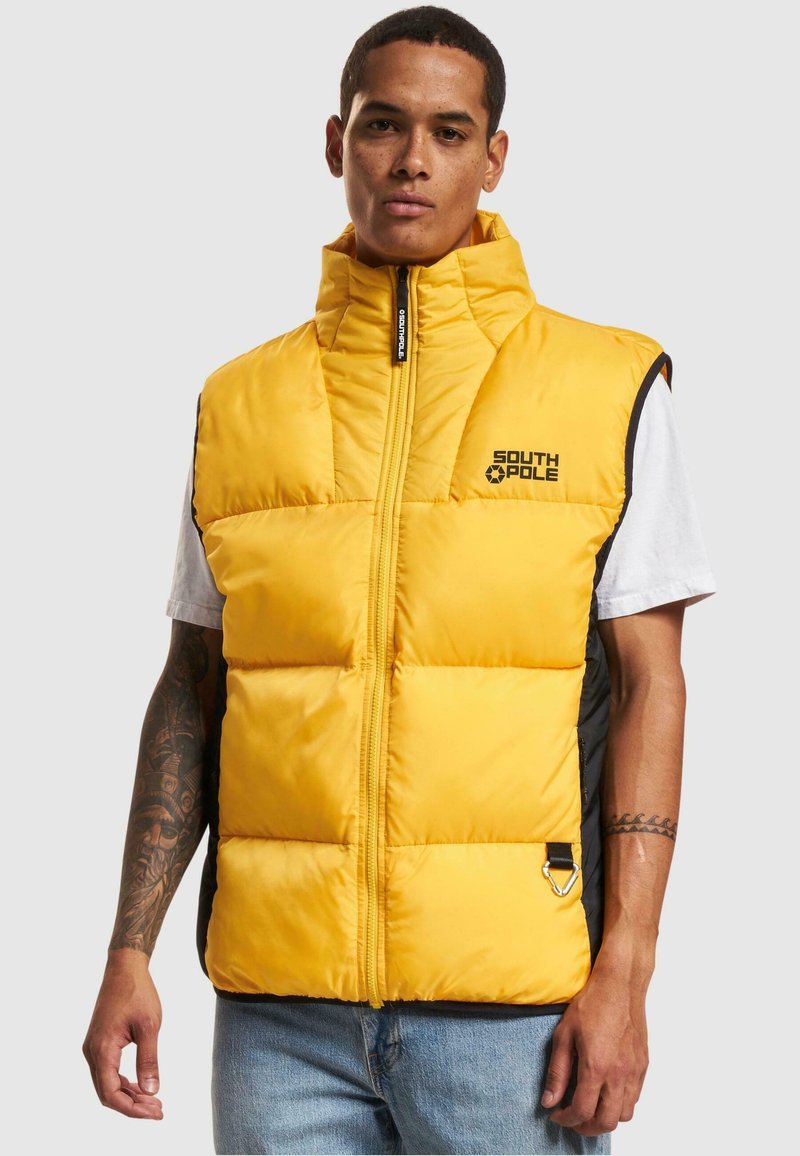 Žltý vypchávkovaný vest s vysokým golierom a čiernymi akcentami. Obsahuje predný zips a logo. Hladká textura, prešívaný dizajn a bočný detail s kovovými prvkami.