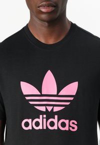 Svart bomull t-skjorte med et stort rosa Adidas-logo med et design med tre blader og tre horisontale striper under logoen.