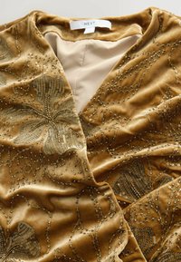 Velvet omslagskjole i guld med detaljeret blomsterperlesyning og en glat indvendig foring. Blød tekstur med en diskret glans.
