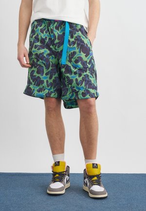 Mężczyzna w zielono-czarnych szortach moro z niebieskim sznurkiem, białych skarpetkach i białych szaro-żółtych sneakersach typu high-top stojący na niebieskim dywanie.