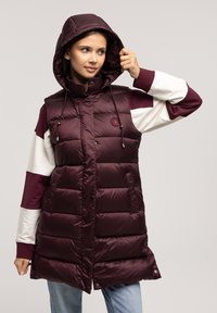 Burgundowa kamizelka puchowa z kapturem, z pikowanym wzorem, detalami sznurków i naszywką z logo. Noszona na kolorowej, blokowanej bluzie z długim rękawem.