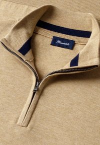 Pull beige à col montant zippé au quart, avec des accents bleu marine à l'intérieur du col et une étiquette de marque bleu marine cousue à l'arrière du col.
