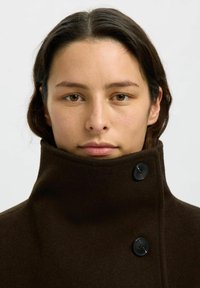 Cappotto di lana marrone con colletto alto, caratterizzato da due bottoni neri sul davanti e una texture liscia. Design semplice e pulito.