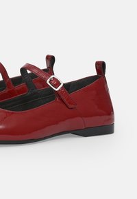 Ballerines Mary Jane rouges brillantes avec doubles brides et boucles argentées, doublure intérieure noire, petits talons noirs et languettes à l’arrière.
