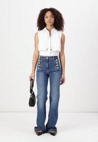 Top bianco corto con pieghe frontali, abbinato a jeans blu a vita alta svasati con dettagli in oro sui bottoni. Borsa nera e scarpe a punta.