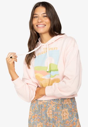 Felpa rosa con un grafiche che presenta un tramonto e un design di paesaggio in colori pastello. Il modello indossa una gonna con fantasia e tiene il cordino della felpa.
