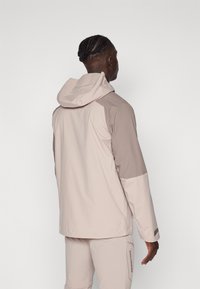 Lightweight, water-resistant jacket in beige and taupe, featuring a hood, contrasting sleeves, and subtle branding on the side.