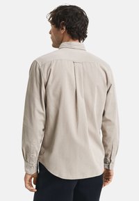 Man in een beige corduroy overhemd met lange mouwen en donkere broek, gezien van achteren tegen een effen witte achtergrond.