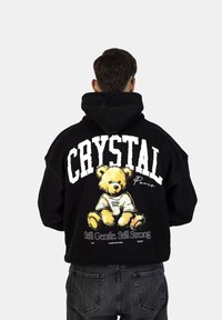 Felpa nera con cappuccio caratterizzata da una grande grafica di un orsetto, il testo "CRYSTAL Paris" e lo slogan "Still Gentle. Still Strong." sul retro.