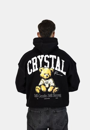 Schwarzer Hoodie mit einem großen Grafikmotiv eines Teddybären, dem Text "CRYSTAL Paris" und dem Slogan "Still Gentle. Still Strong." auf dem Rücken.