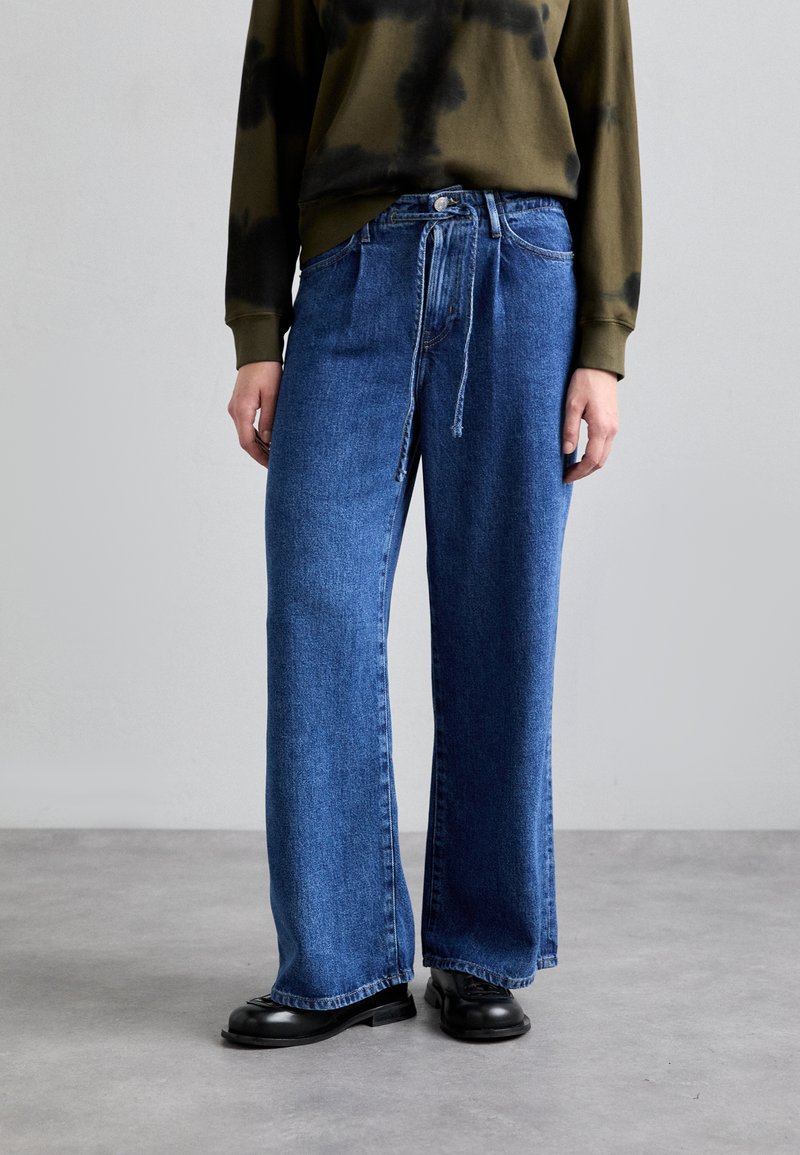 Jeans din denim albastru cu picioare largi și talie cu sfoară, prezentând un finisaj decolorat și o textură subtilă. Asortate cu un pulover dark tie-dye și cizme negre.